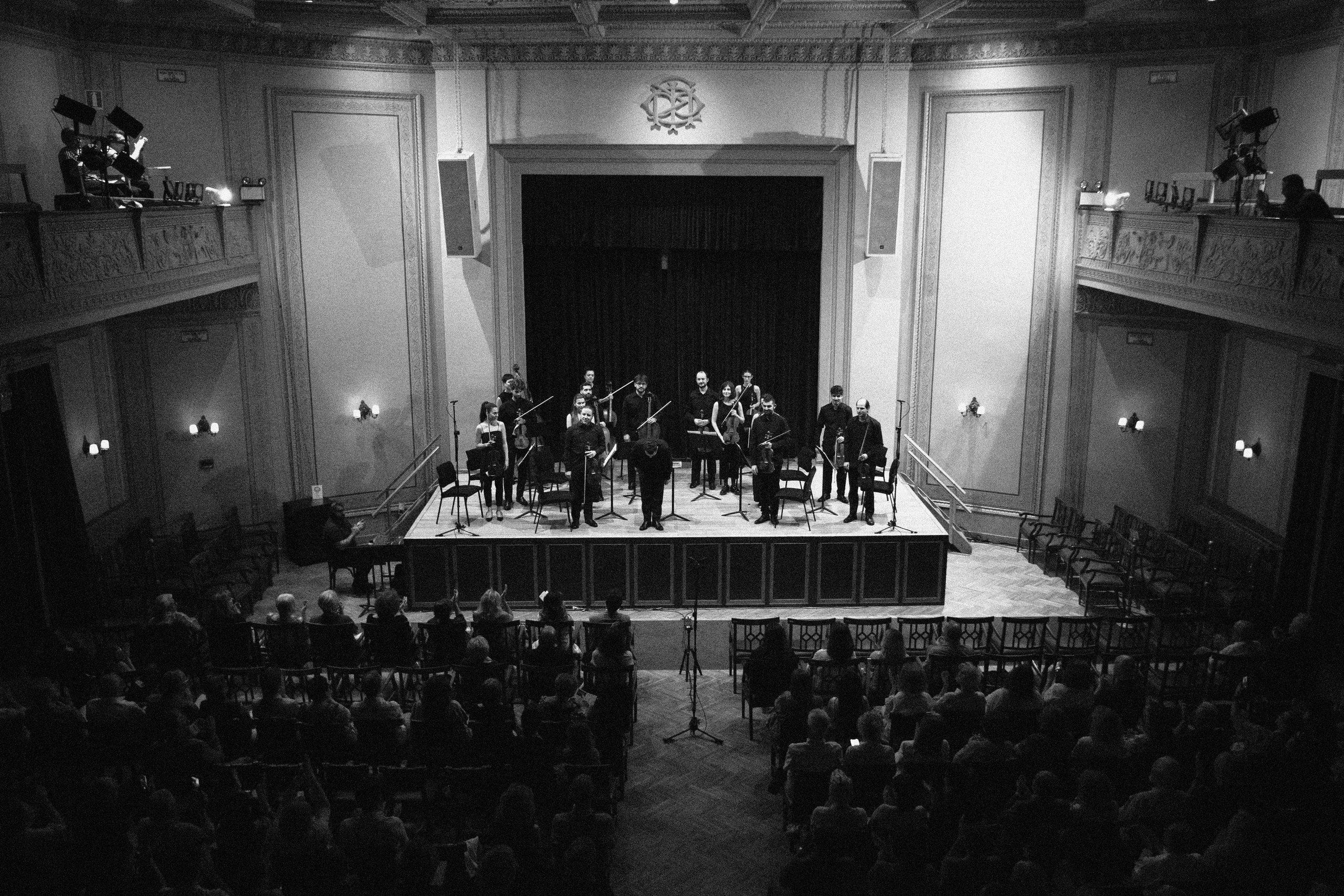 The ensemble on a lit stage at the far end of a high, ornate concert hall, seen over the heads of a full audience.